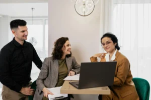 Three people in business attire engage in a meeting. A woman in a brown blazer sits at a laptop, while the others smile, creating a collaborative atmosphere.